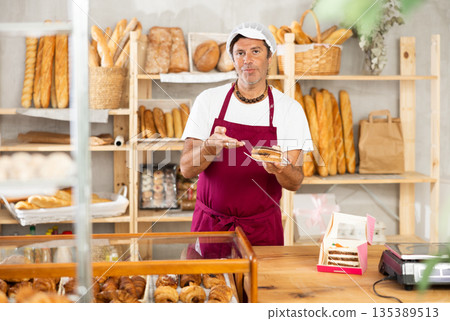 Middle-aged salesman offering a piece of cake in plate in bakery 135389513