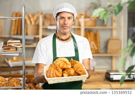 Portrait of a happy male baker offering to buy fresh delicious croissants in bakery Portrait of a happy male baker offering to buy fresh delicious croissants in bakery 135389552