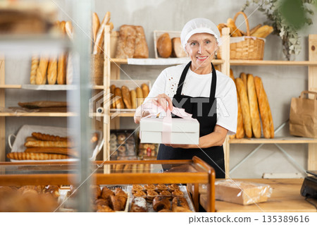 Senior woman entrepreneur owner of bakery holds out cardboard box with pastries in hand Senior woman entrepreneur owner of bakery holds out cardboard box with pastries in hand 135389616