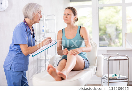 Mature female doctor listens to complaints of knee pain of woman before procedure and fill in report Mature female doctor listens to complaints of knee pain of woman before procedure and fill in report 135389655