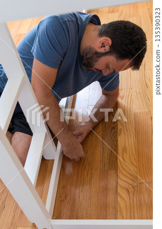 A man is kneeling on the floor and fixing a piece of furniture A man is kneeling on the floor and fixing a piece of furniture 135390323
