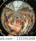 Fisheye perspective of a bustling shopping mall with a hand in the foreground. Fisheye lens captures a busy shopping mall scene, featuring a transparent arched ceiling allowing natural light. 135391048