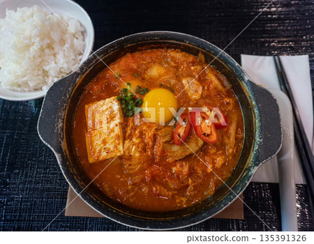 Authentic Korean Kimchi Jjigae or spicy kimchi stew with tofu, egg yolk, and chili in a hot pot served with steamed rice. Traditional Asian comfort food photography 135391326