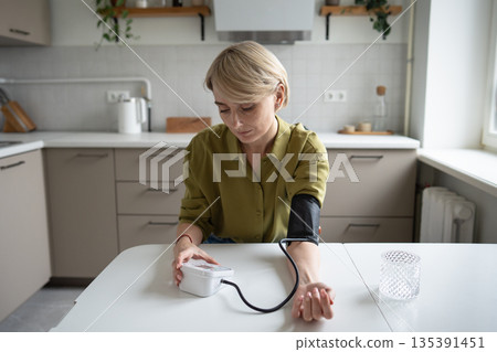 Middle aged woman sits at table measuring blood pressure with tonometer, seasonal health problems. 135391451