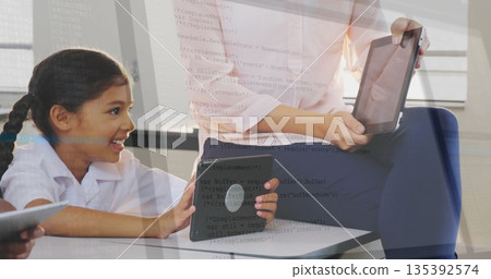 Holding tablet teacher guiding girl in collared shirt leaning at classroom desk, with second tablet 135392574