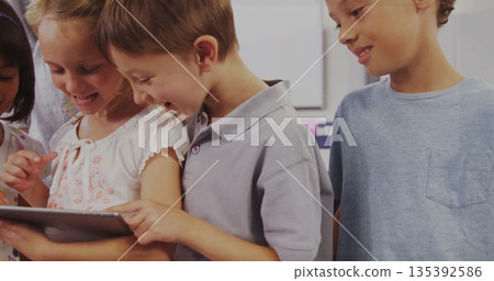 Girl in white floral top holding tablet with peers tapping tablet in classroom, desks whiteboard 135392586