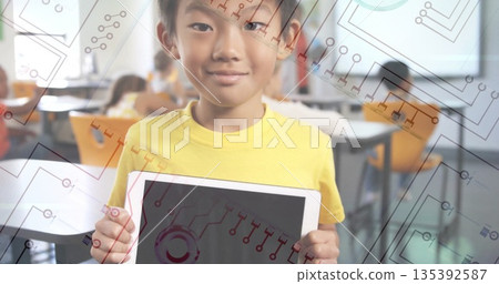 Standing boy holding tablet displaying circuit-pattern graphics in classroom, with orange seats Standing boy holding tablet displaying circuit-pattern graphics in classroom, with orange seats 135392587