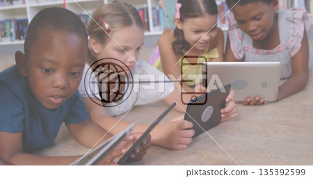 Lying four children wearing colorful clothes using tablets in reading area, with bookshelves behind 135392599