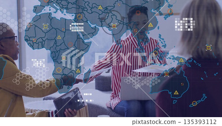 Hispanic woman in blouse exchanging documents on laptop in office lounge with world map projection 135393112