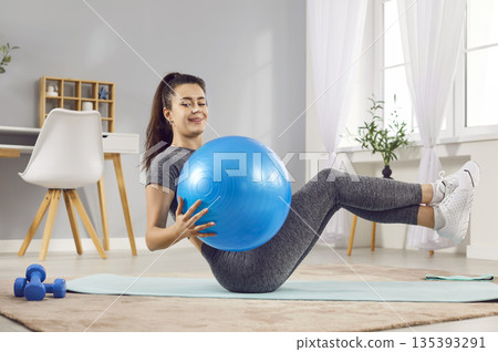 Young cheerful girl doing fit exercises lying on yoga mat using fit ball at home. Young cheerful girl doing fit exercises lying on yoga mat using fit ball at home. 135393291