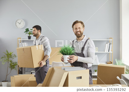 Smiling mover holding office plant while coworker carries boxes during relocation 135393292