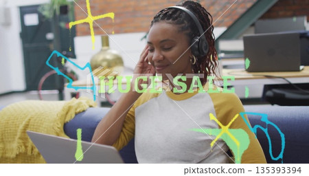 Typing Black woman wearing headphones on couch in office, working on laptop with neon sale text 135393394