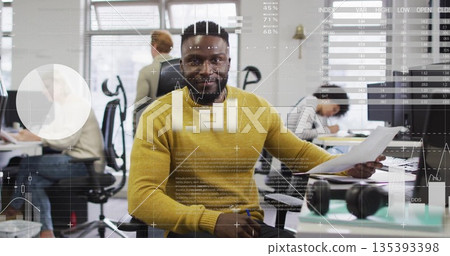 Man in mustard-yellow sweater analyzing documents at open-plan office, with data overlays 135393398