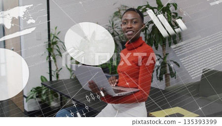 Holding laptop, woman wearing red turtleneck presenting charts in office lounge with world map 135393399