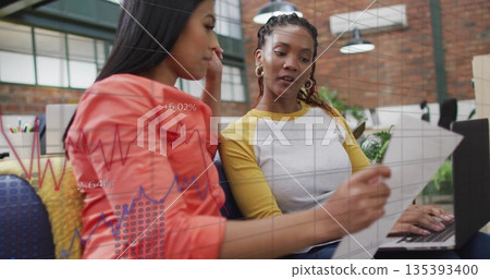 Reviewing colleagues working on laptop and printed document in office lounge, showing data overlay 135393400