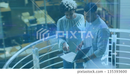 Standing coworkers pointing at laptop on mezzanine with metal railing above desks shelving units 135393600