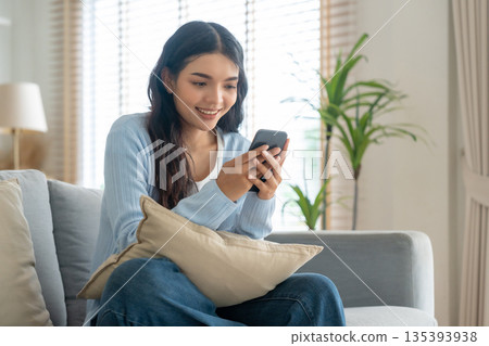 Smiling pretty young Asian woman sitting on cozy couch Smiling pretty young Asian woman sitting on cozy couch 135393938