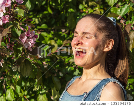 Young woman sneezes in the park against the background of a flowering tree. Allergy to pollen concept. Young woman sneezes in the park against the background of a flowering tree. Allergy to pollen concept. 135394176