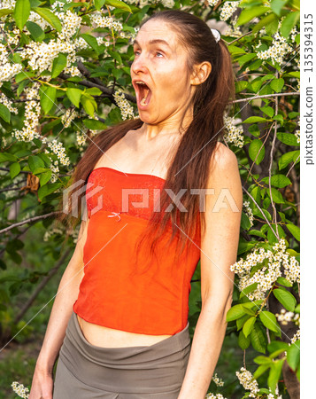 Young woman sneezes in the park against the background of a flowering tree. Allergy to pollen concept. 135394315