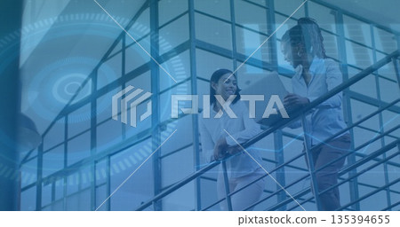 Standing two colleagues discussing in office atrium, with laptop and digital interface graphics Standing two colleagues discussing in office atrium, with laptop and digital interface graphics 135394655