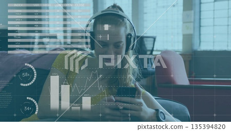 Using smartphone, woman wearing yellow top, headphones, fitness watch in lounge, with stock overlay 135394820