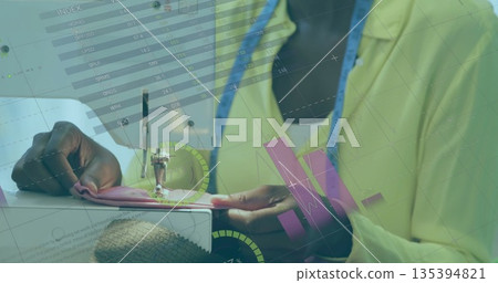 Seamstress guiding pink fabric under sewing machine in studio, with measuring tape and charts Seamstress guiding pink fabric under sewing machine in studio, with measuring tape and charts 135394821