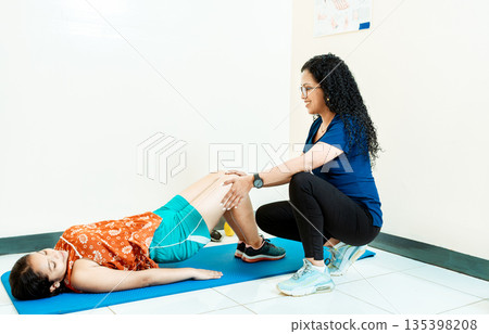 Woman physiotherapist assisting patient lying down doing exercises on the mat, Physiotherapist with patient on the floor doing knee rehabilitation exercises Woman physiotherapist assisting patient lying down doing exercises on the mat, Physiotherapist with patient on the floor doing knee rehabilitation exercises 135398208