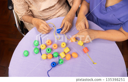 Caregiver and senior woman playing stringing beads game for dementia prevention 135398251
