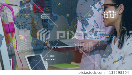 Standing colleagues wearing office attire, viewing laptop screen at office with world map overlay 135398304