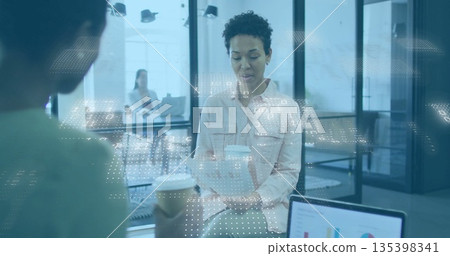 African woman holding takeaway cup and reading printed document in office area, with laptop charts 135398341