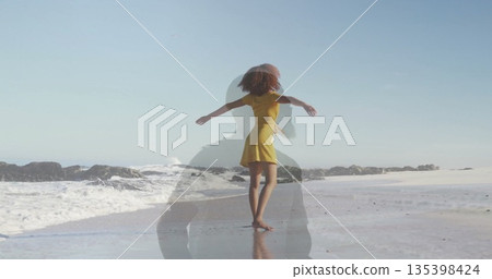 Stretching woman in yellow dress facing ocean at beach, with wet sand, ocean waves, rocky outcrop 135398424