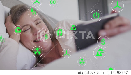 Lying senior woman holding tablet, smiling at screen in bedroom on bedding with eco-icons overlay Lying senior woman holding tablet, smiling at screen in bedroom on bedding with eco-icons overlay 135398597