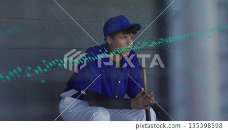 Sitting teen in blue uniform and cap gripping wooden bat on dugout bench, gazing toward field 135398598