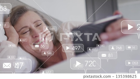 Relaxing mature woman holding tablet while reclining in bedroom, with pillows and bed linens 135398625