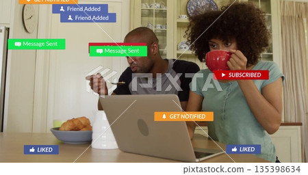 Sitting couple using silver laptop at kitchen table, with red mug, cereal bowl and notifications 135398634