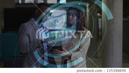 Two colleagues wearing suits reviewing tablet in office corridor, with holographic security overlay Two colleagues wearing suits reviewing tablet in office corridor, with holographic security overlay 135398640