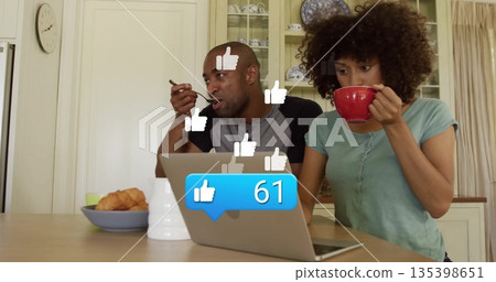 Mid adult couple eating breakfast while using silver laptop at wooden kitchen table, with red mug 135398651