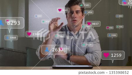 Man wearing gray shirt sitting at home office interacting with tablet, floating notification icons 135398652