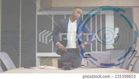 Standing man in navy suit behind table in boardroom, with notebook pen speakerphone laptop 135398653