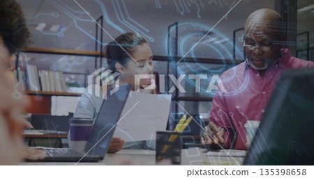 Collaborating woman and senior man examining document at conference table with laptops, overlays 135398658