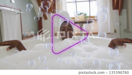 Woman with towel-wrapped hair soaking in foam-filled white bathtub in bathroom, with neon play icon Woman with towel-wrapped hair soaking in foam-filled white bathtub in bathroom, with neon play icon 135398760
