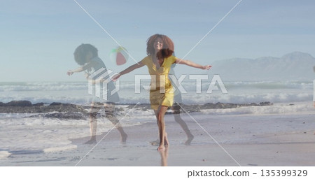 Twirling woman wearing yellow dress walking along wet sand at ocean edge, with floating beach ball Twirling woman wearing yellow dress walking along wet sand at ocean edge, with floating beach ball 135399329
