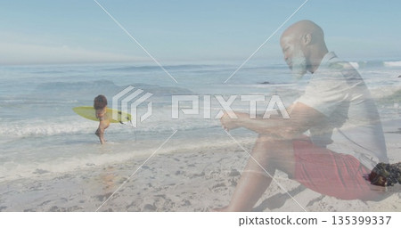 Child in swimsuit carrying surfboard wading at shoreline with senior checking smartphone, beach bag 135399337