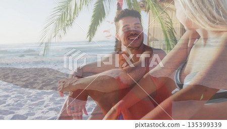 Smiling friends in swimwear relaxing on white sand beach under palm tree frond, ocean waves behind Smiling friends in swimwear relaxing on white sand beach under palm tree frond, ocean waves behind 135399339