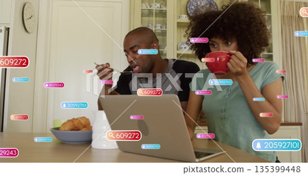 Man and woman eating and drinking at home kitchen table, with laptop, red mug and notifications Man and woman eating and drinking at home kitchen table, with laptop, red mug and notifications 135399448