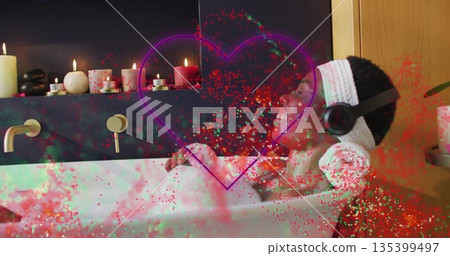 Reclining woman wearing towel listening through headphones in tub with foam pillow and lit candles 135399497