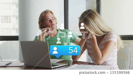 Collaborating couple discussing documents at office table, with laptop and notification graphics 135399566