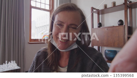 Smiling mature woman leaning forward in softly lit room, with mask under chin and chess set 135399568