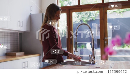 Woman in sweatshirt washing hands under high-arc faucet into sink in home kitchen with social icons 135399652