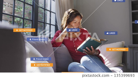 Woman wearing red top, blue jeans, reading book on sofa in living room, with notification icons 135399693
